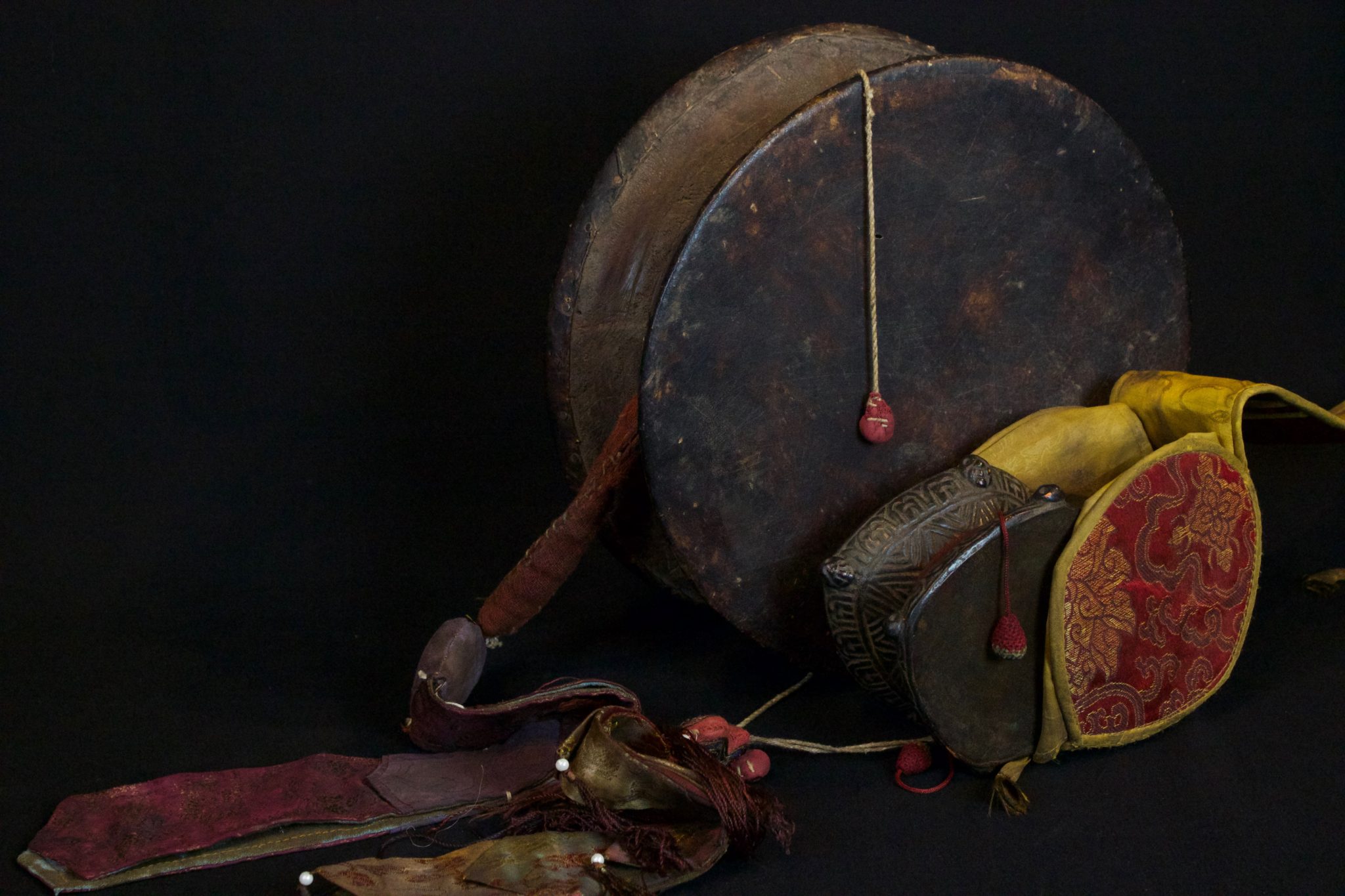 Damaru with Chopin (2 sided drum with sash), late 19th and early 20th c, wood, hide, fabric This power drum, associated with Shiva, is used for tantric rituals and is played with a twisting motion so the two beaters - attached by cords, hit the drum heads simultaneously. It produces a spiritual sound by which the universe was created and is regulated. The Chopin is typically embroidered with the colors of tantric elements and waves as the drum is played. large - 32" x 11" x 4 ", $700.; small with case